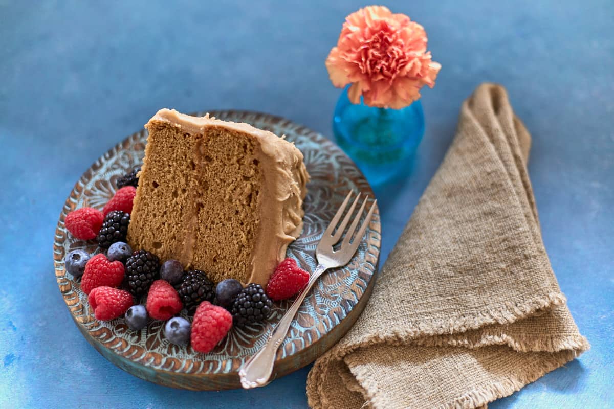 a full slice of cake on a plate surrounded by berries with a fork, and a napkin on the side and a flower in a vase in the background