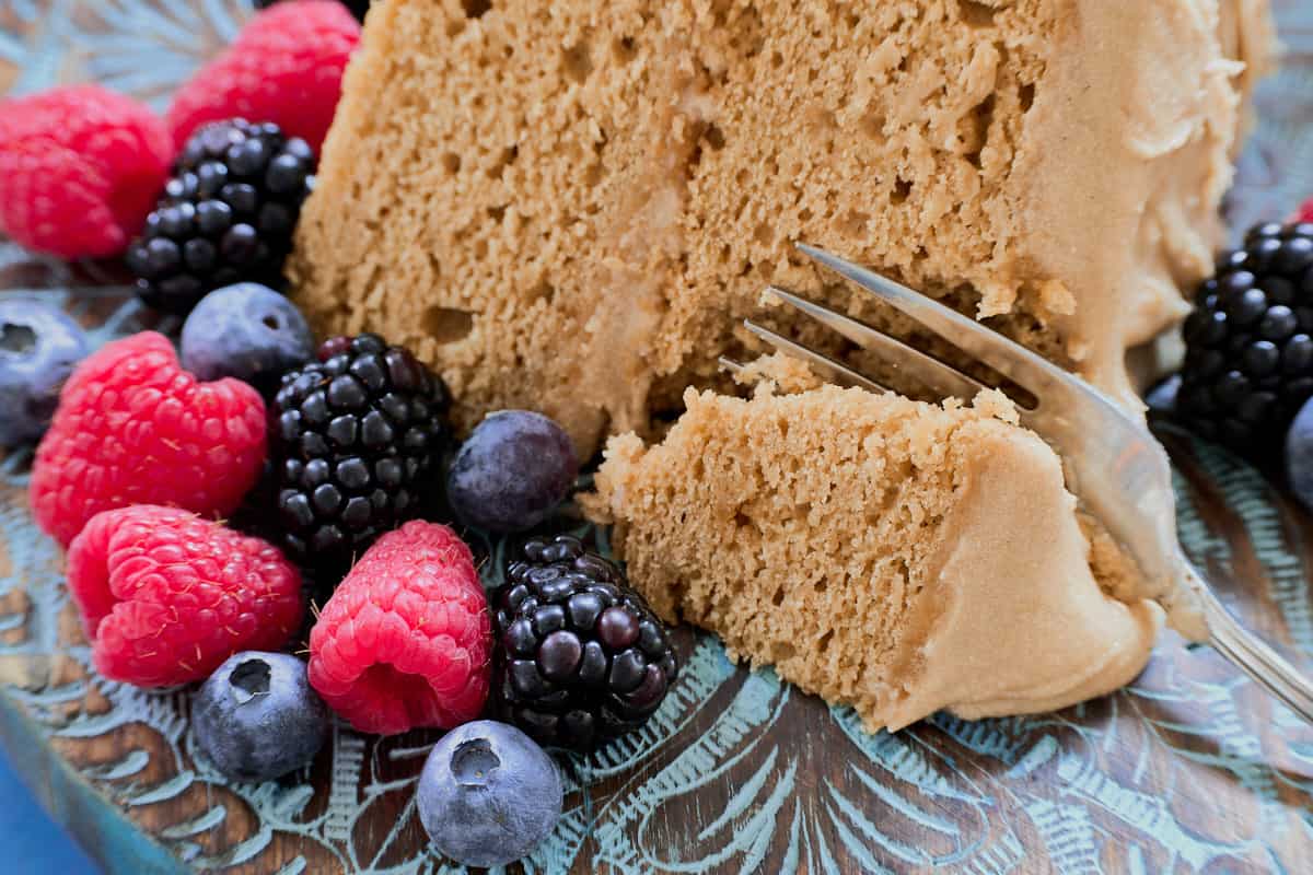a close-up shot of a slice of cake on a wooden plate surrounded by berries, with a fork removing the first bite from the slice
