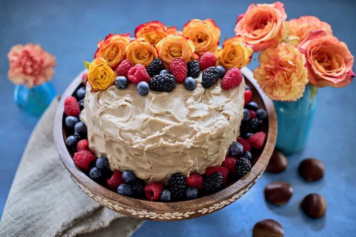 cake decorated with berries and flowers on a stand, with vases of flowers in the background and a napkin and chestnuts in the foreground