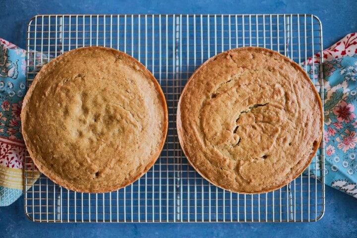two 8 inch freshly baked cakes , just out of the oven on a cooling rack with kitchen towels on the sides of the rack