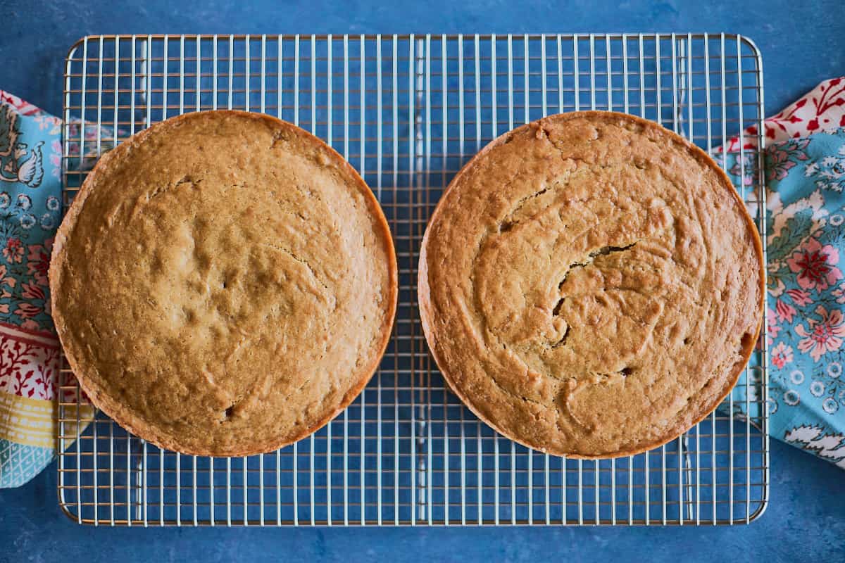 two 8 inch freshly baked cakes , just out of the oven on a cooling rack with kitchen towels on the sides of the rack