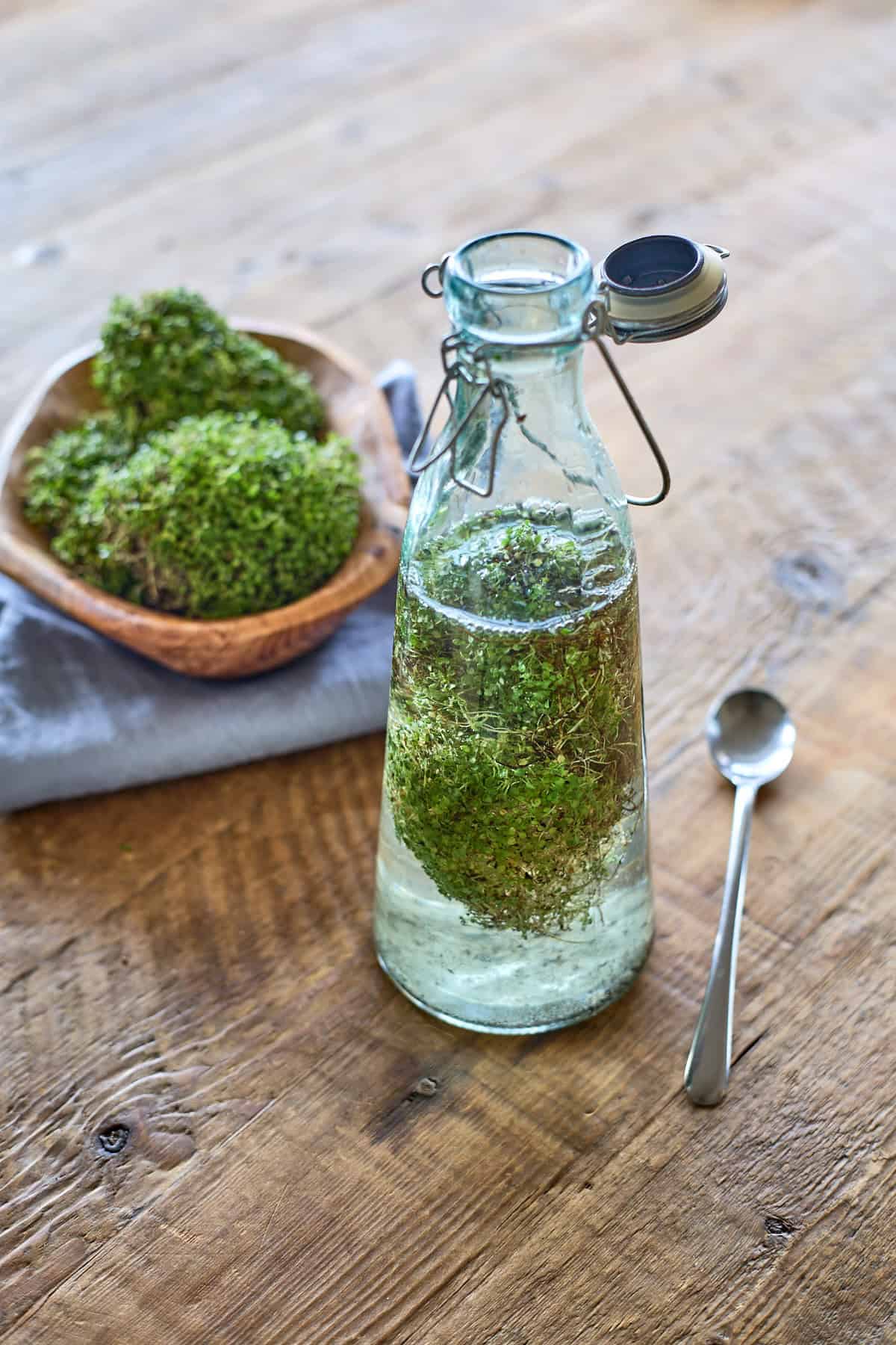 a bottle of vodka with fresh corsican mint leaves, with a wooden bowl of mint on a napkin in the background, and a metal spoon on the table alongside the bottle