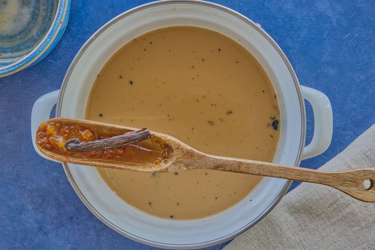 a white pot filled with chai and a wooden cooking spoon balanced atop the pot with a piece of vanilla bean and some wet jaggery (raw cane sugar) chunks in the spoon, with a kitchen towel and a blue ceramic bowl in the background