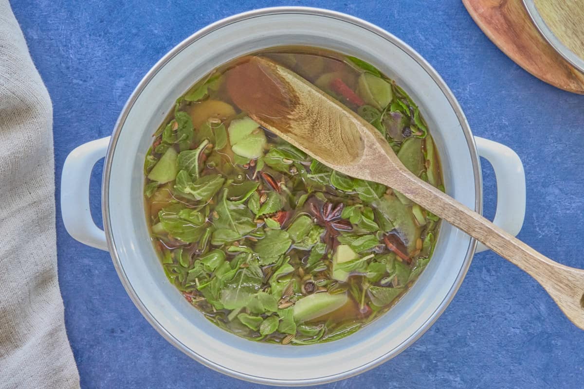 a white pot filled with water, spices, and tulsi (holy basil) leaves, with a wooden spoon in the pot, and a kitchen towel, wooden cutting board, and pot lid in the background