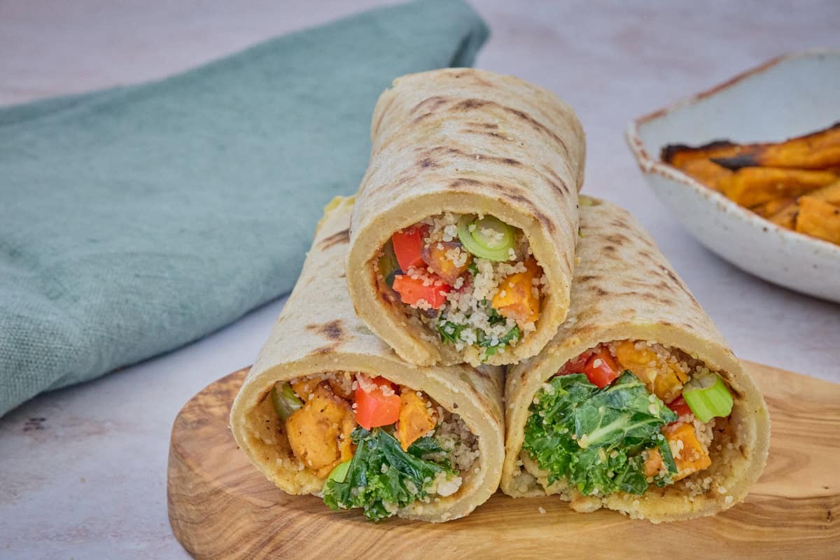 three filled wraps on a wooden board with a napkin and dish of roasted sweet potatoes in the background