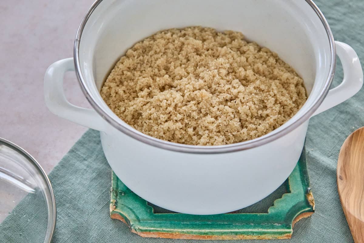 a pot containing cooked fonio on a trivet resting on a napkin, with the pot lid and a wooden cooking spoon alongside