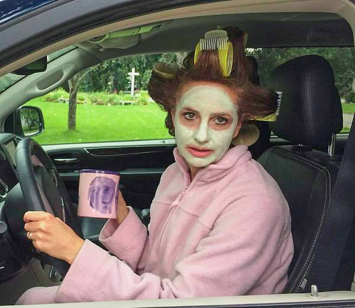 the author sitting in a car in a bathrobe, curlers, facial mask, holding a coffee, looking disdainful