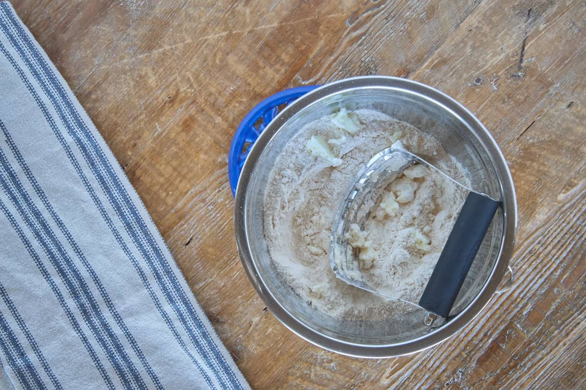 a bowl of dough ingredients with a pastry cutter cutting chunks of butter into the dough, and a kitchen towel alongside