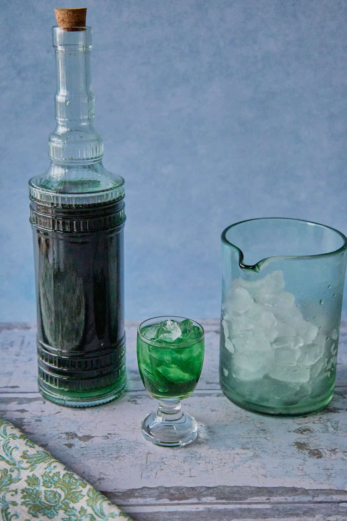 a bottle of liqueur, a mixing glass with ice, and a small glass full of liqueur and ice, with a napkin in the foreground