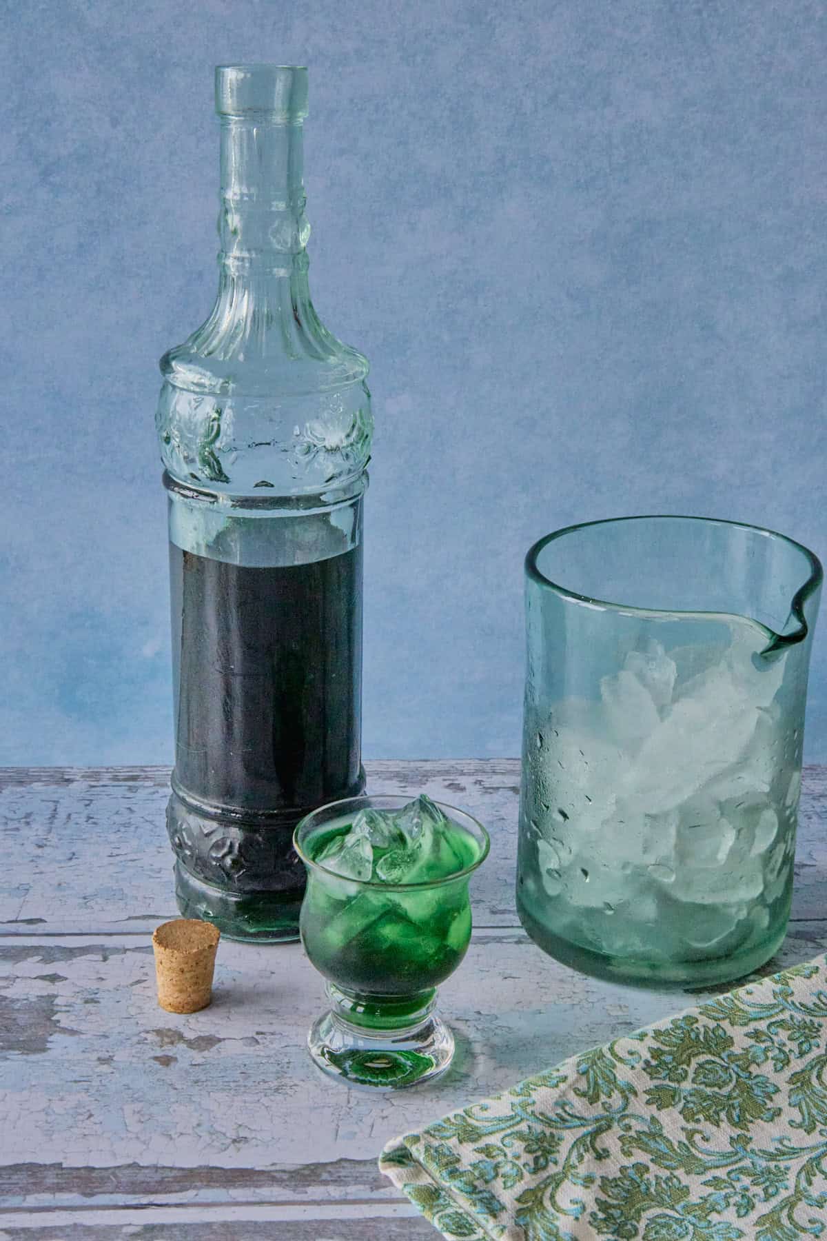 a bottle of liqueur, a mixing glass with ice, and a small glass full of liqueur and ice, with a napkin in the foreground