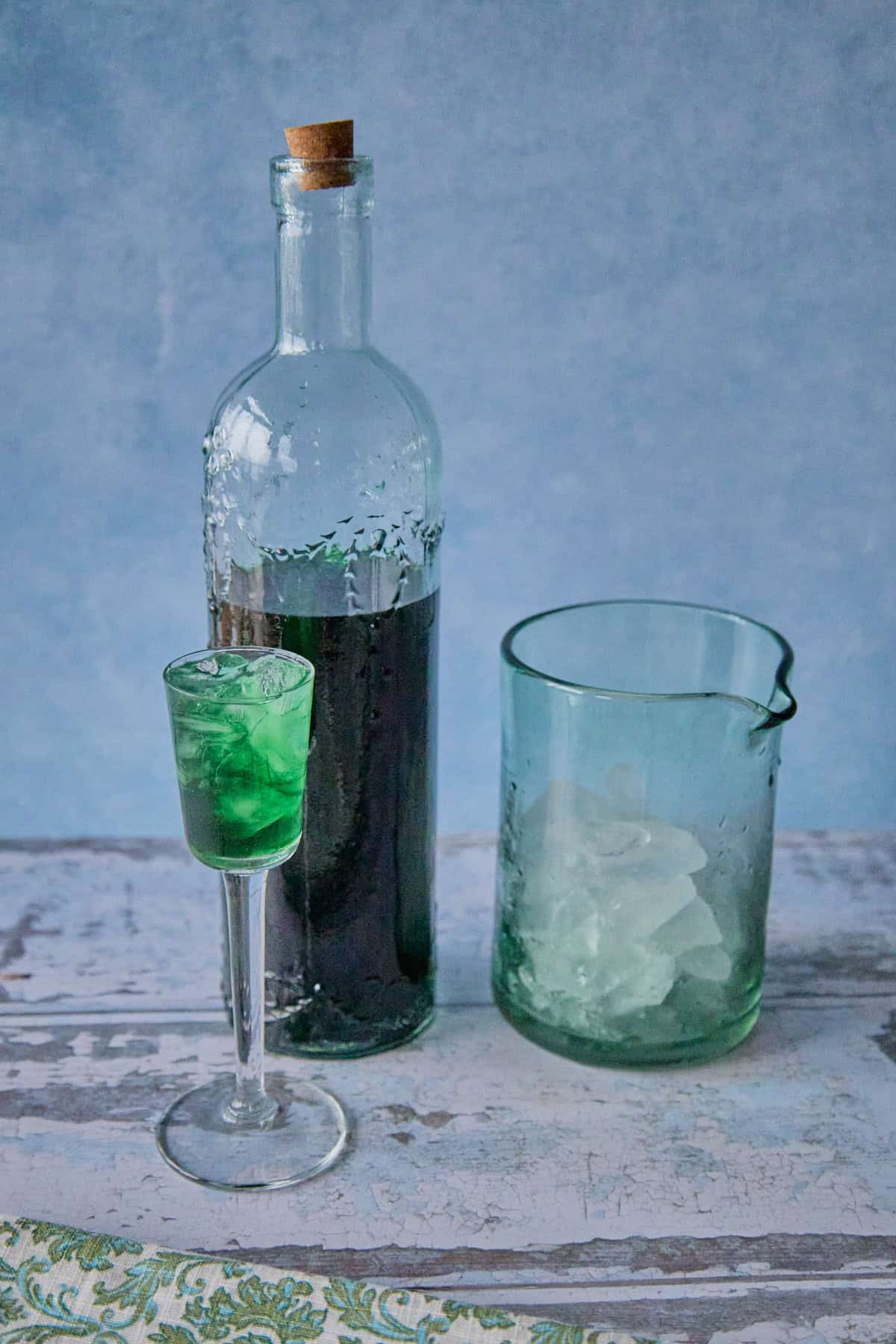 a bottle of liqueur, a mixing glass with ice, and a small glass full of liqueur and ice, with a napkin in the foreground