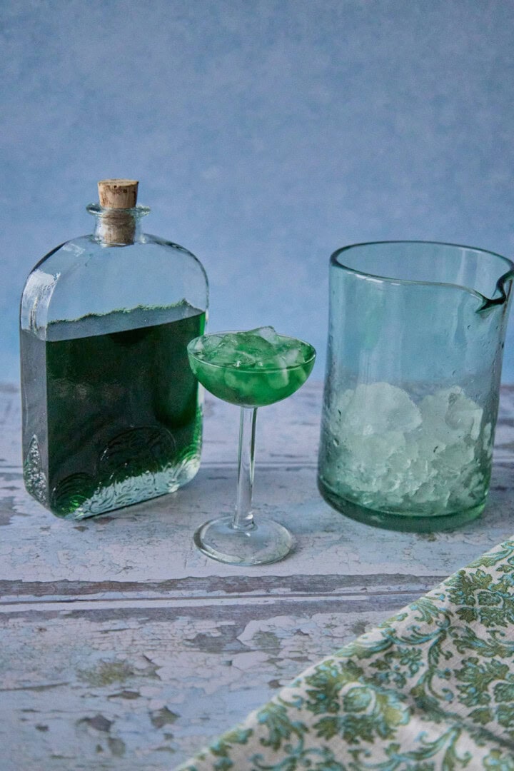 a bottle of liqueur, a mixing glass with ice, and a small glass full of liqueur and ice, with a napkin in the foreground