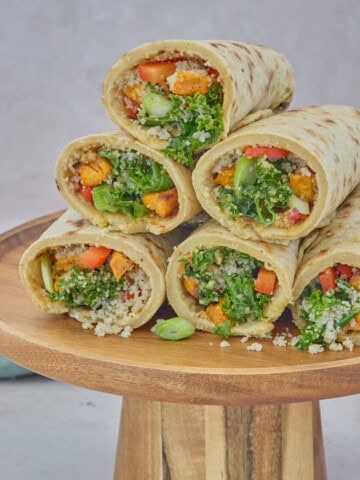 square photo of filled wraps stacked on a wood cake stand, with a napkin and stack on tortillas on a plate in the background