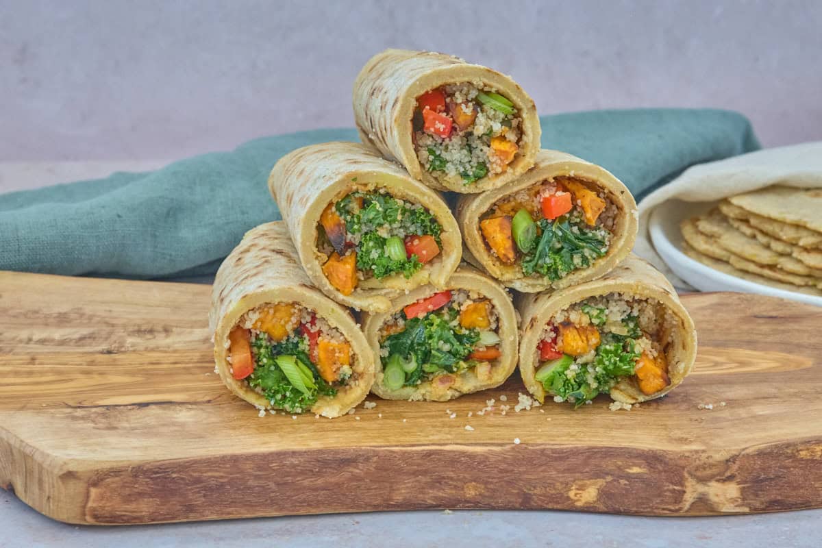 stacked filled wraps atop a wooden board, with a napkin and plate of stacked tortillas topped by a kitchen towel in the background