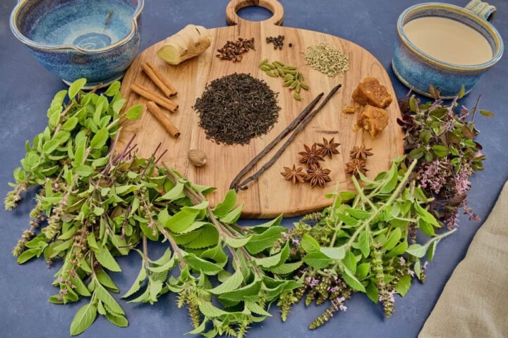 a head-on shot of a cutting board filled with small piles of spices and black tea, with a pile of 5 types of fresh tulsi (holy basil) stems and leaves alongside, and a kitchen towel and bowls of water and non-dairy milk in the background