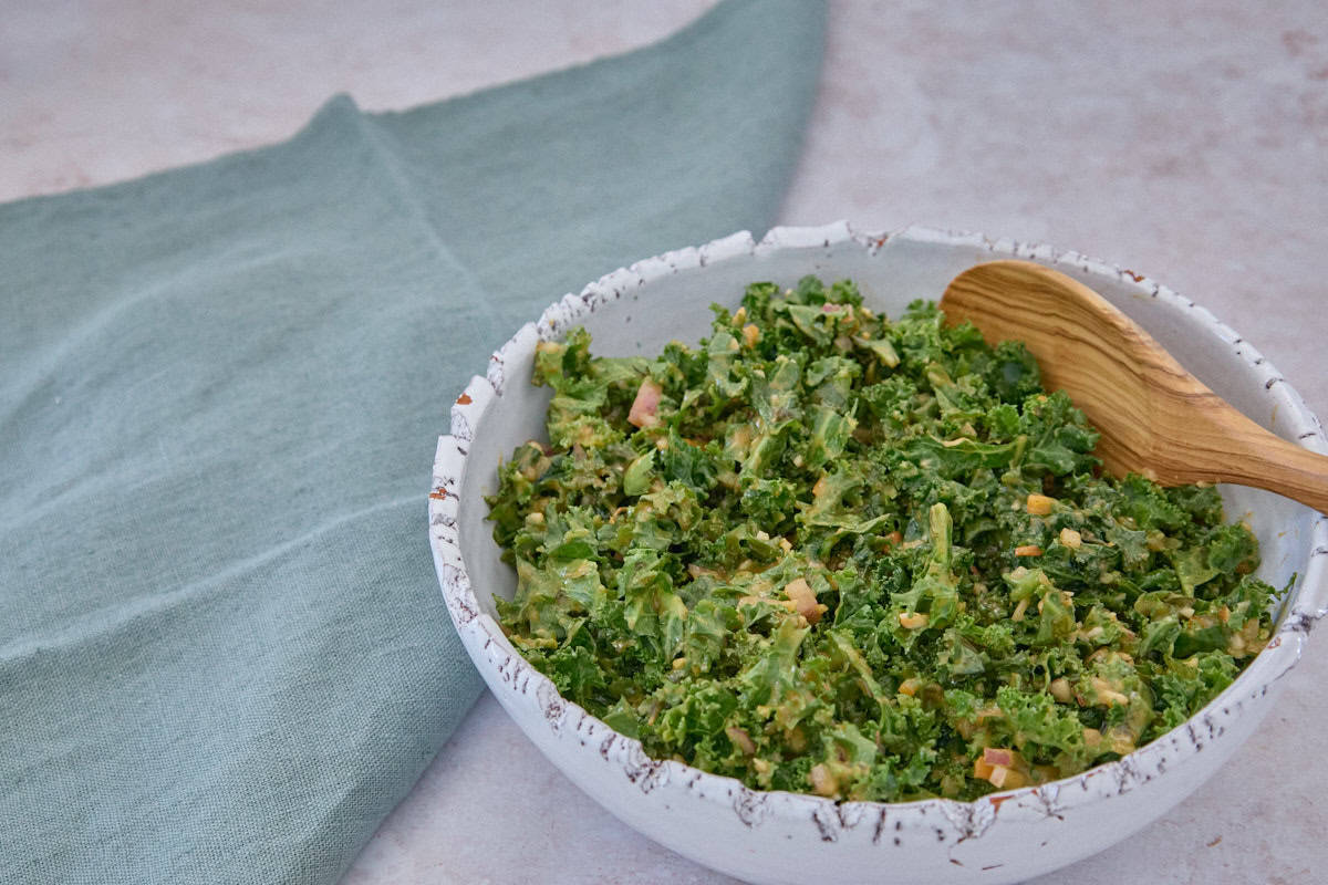 a bowl of kale and mango yassa sauce being stirred together with a wooden spoon, with a napkin alongside