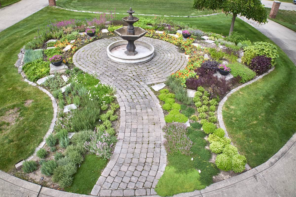 a garden full of herbs bordered by a curved brick edge with an internal path with an empty fountain in the center
