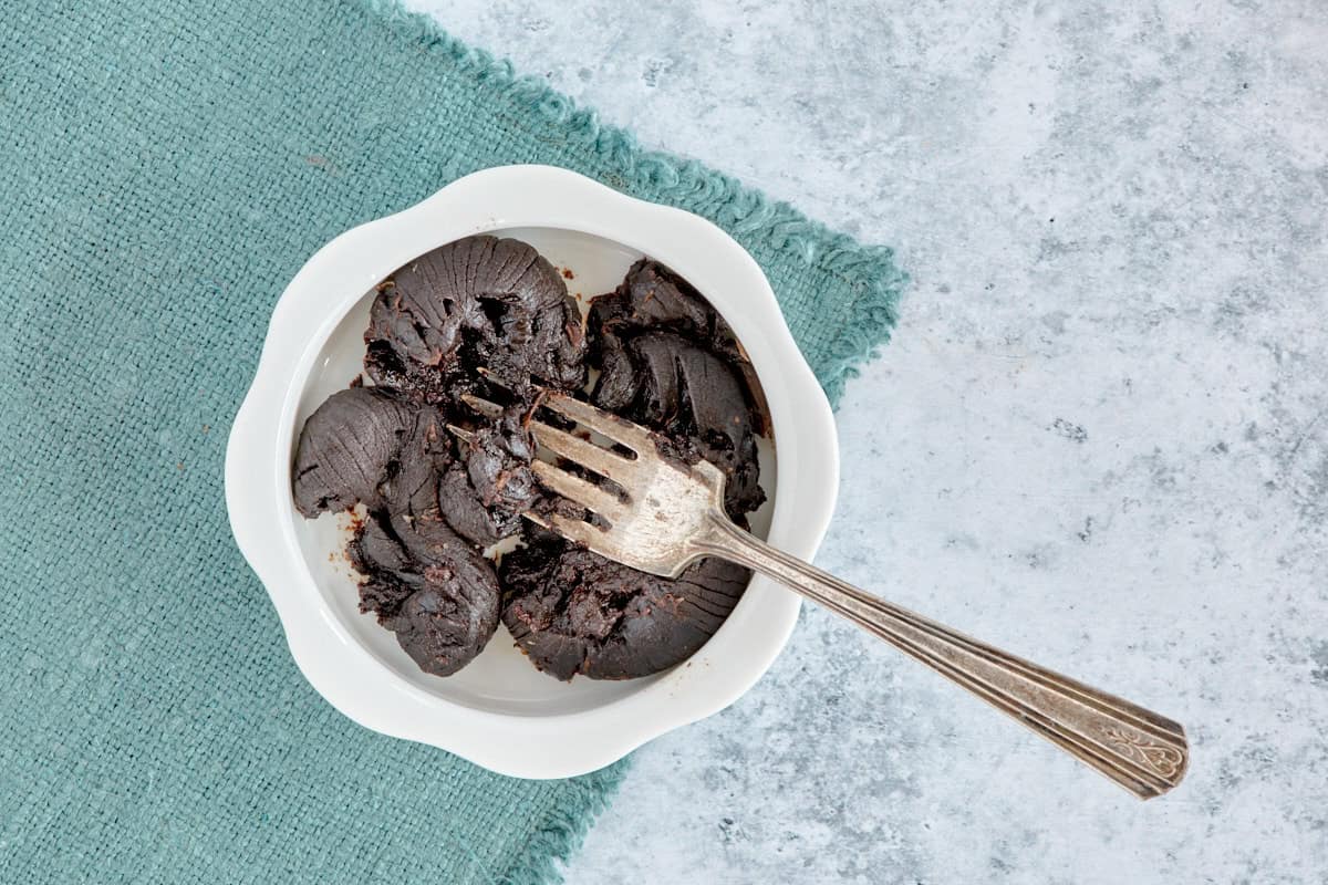 heads of black garlic in a bowl being mashed by a fork, with a napkin beneath