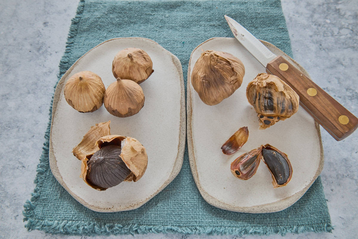 black garlic single-clove heads on a plate on the left and black garlic multiple-clove heads on a plate on the right, with a knife on the plate and a napkin beneath