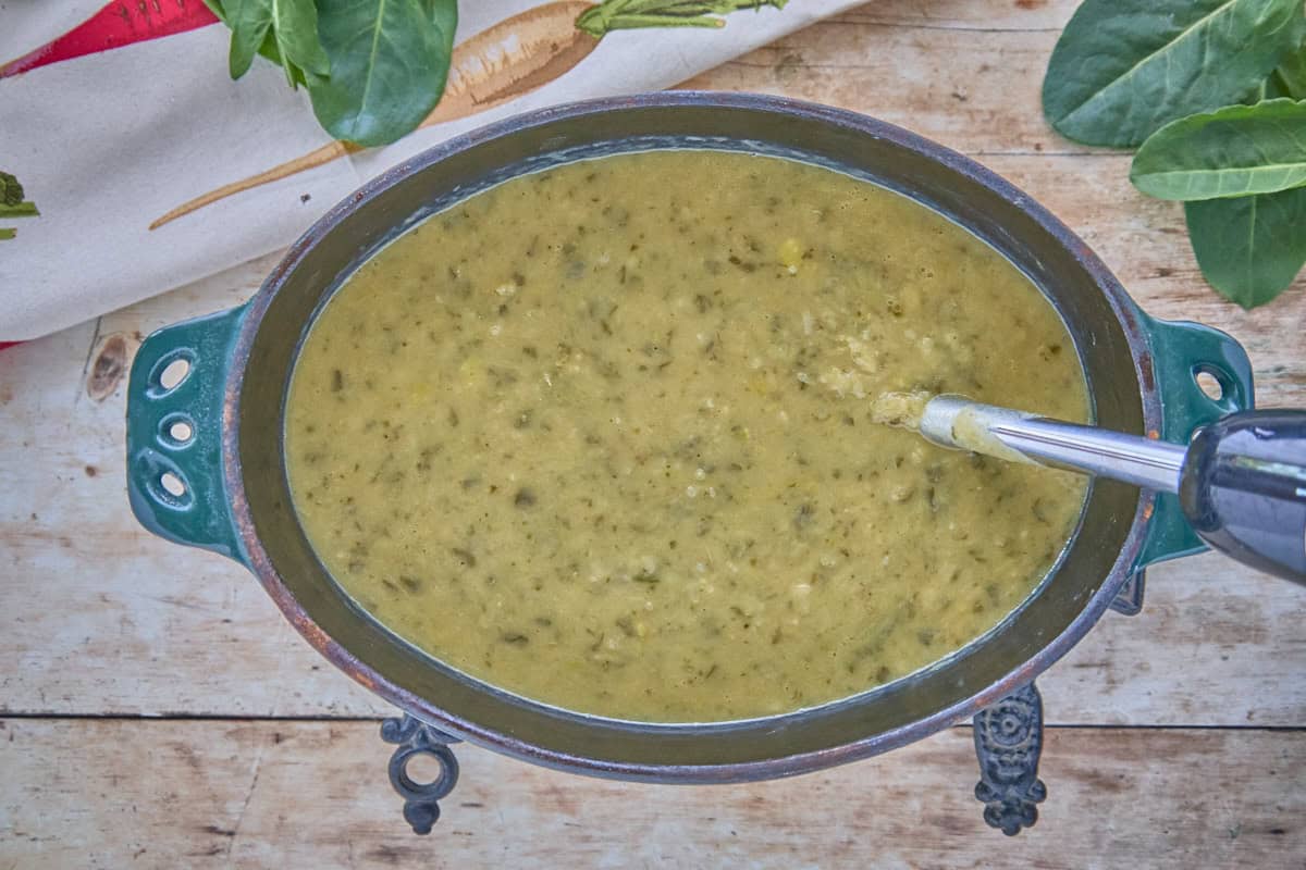 a cast iron pot filled with soup that's being pureed with an immersion blender, with trivets, a kitchen towel, and fresh sorrel leaves in the background
