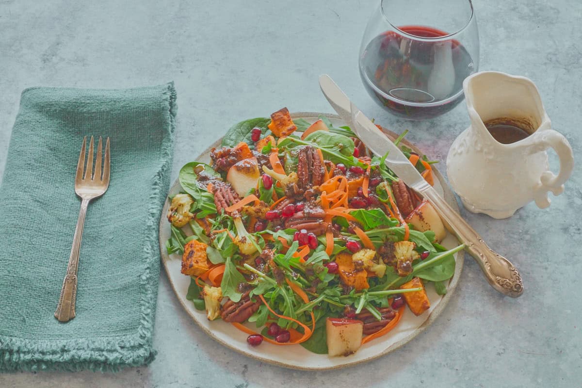 salad on a plate topped with dressing, with a fork, knife, napkin, stemless glass of red wine, and small pitcher of dressing
