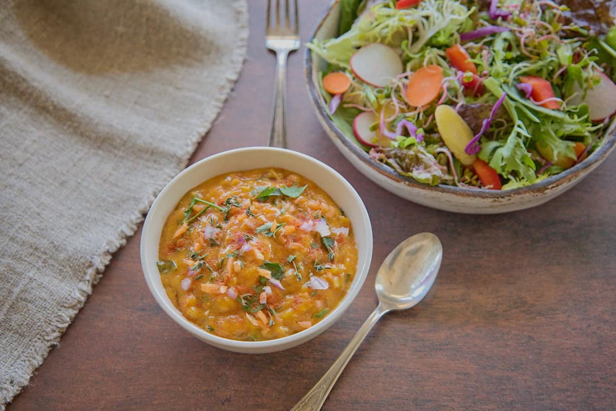 a bowl of sauce in the foreground with a napkin, fork, and spoon, and a larger bowl of undressed salad in the background