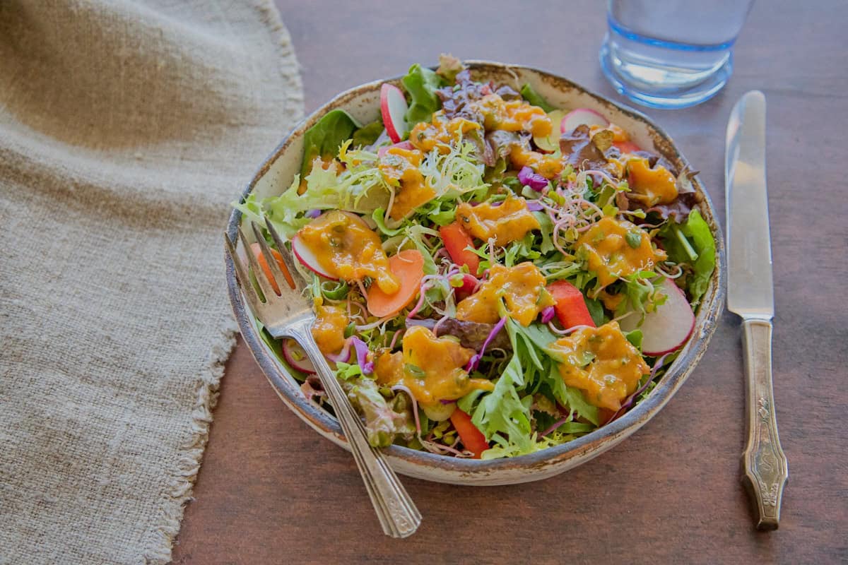 a bowl of salad with sauce on top with a fork, knife, napkin, and glass of water in the background