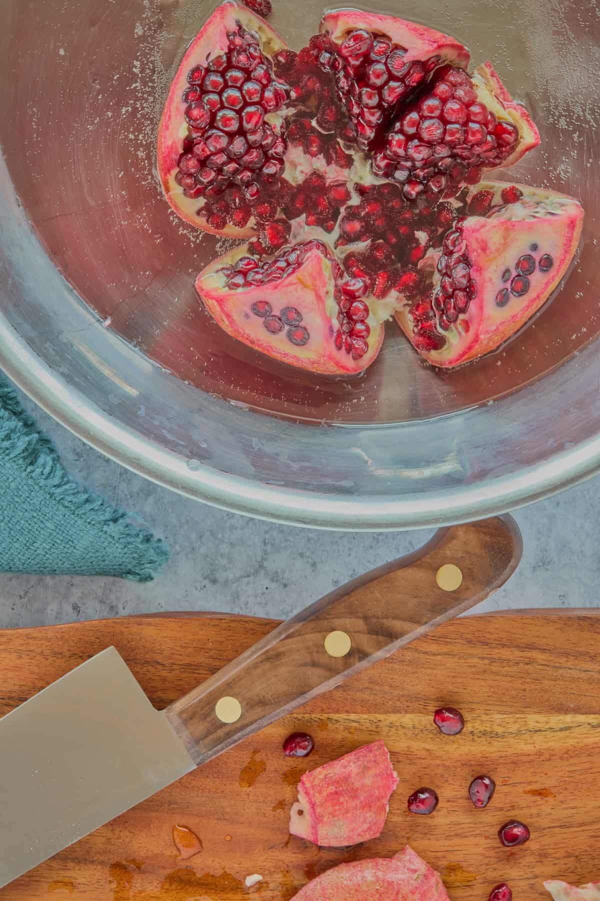 an opened pomegranate in a large bowl of water with a cutting board in the foreground with pomegranate chunks and arils and a knife, and a napkin alongside