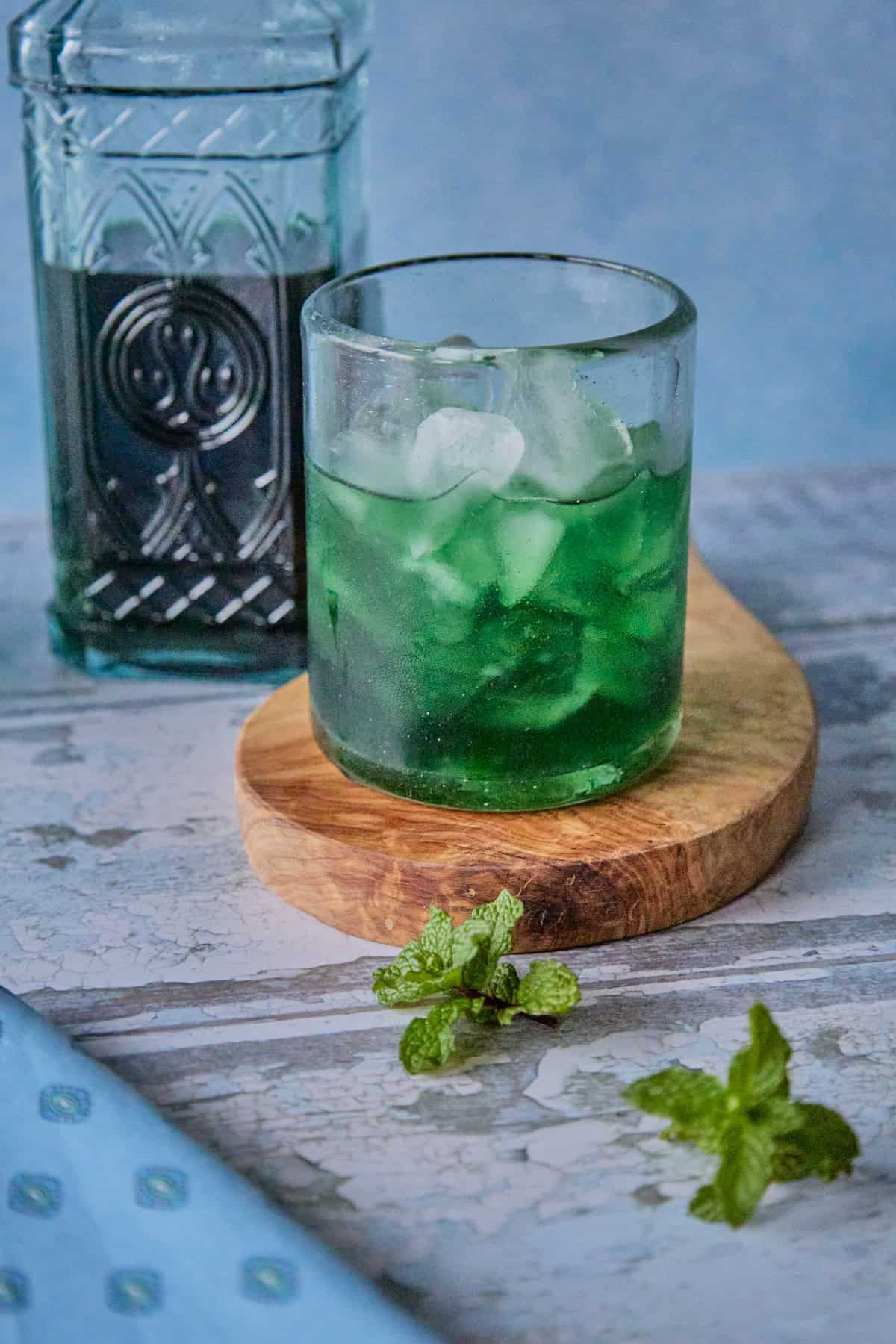 a bottle of liqueur, a small glass full of liqueur and ice on a round wooden board, with a napkin and some fresh mint springs in the foreground