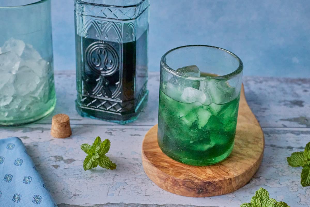 a bottle of liqueur, a cork, a mixing glass with ice, and a small glass full of liqueur and ice on a round wooden board, with a napkin and some fresh mint springs in the foreground
