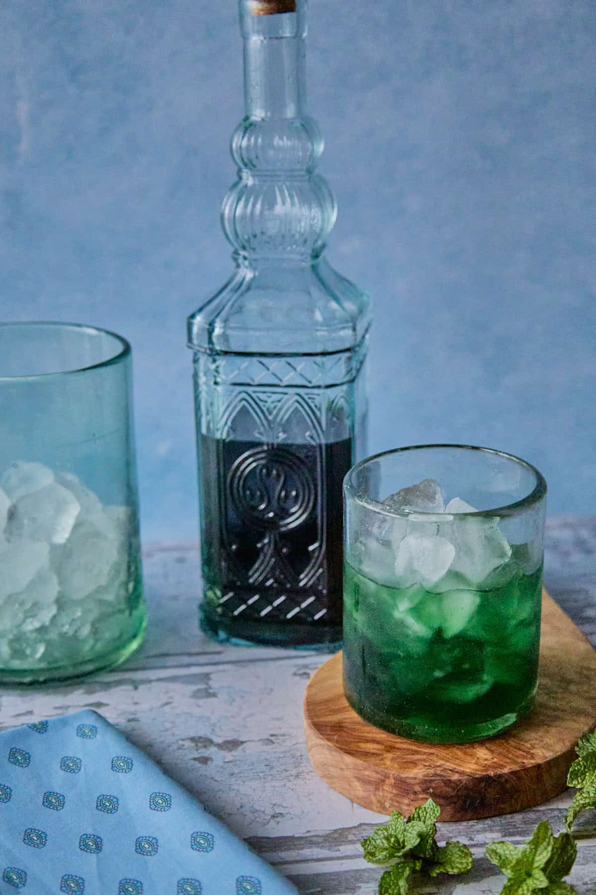 a bottle of liqueur, a mixing glass with ice, and a small glass full of liqueur and ice on a round wooden board, with a napkin and some fresh mint springs in the foreground