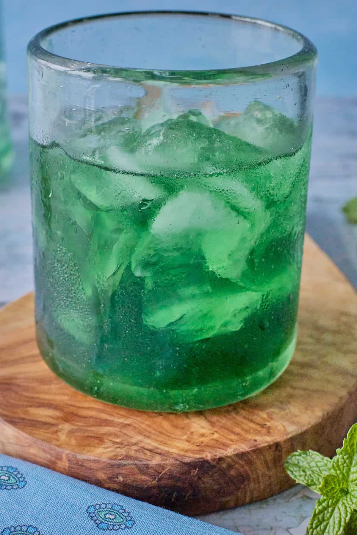 a glass full of liqueur and ice on a wooden board, with a bottle in the background and a napkin and mint sprig in the foreground