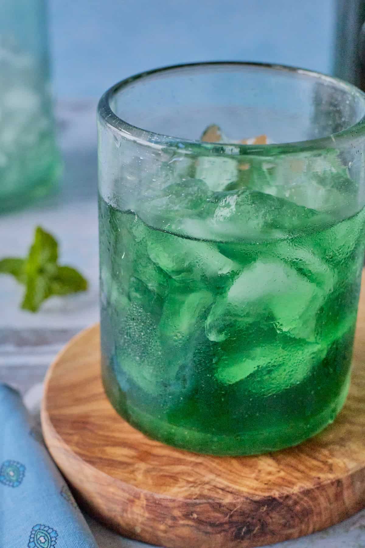 a bottle of liqueur, a mixing glass with ice, and a small glass full of liqueur and ice on a round wooden board, with a napkin and a fresh mint spring in the background