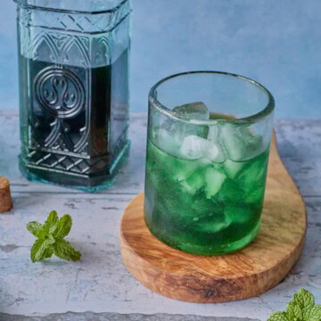 a glass of liqueur and ice on a round wooden board with a bottle of liqueur in the background, and a cork and some mint sprigs in the foreground