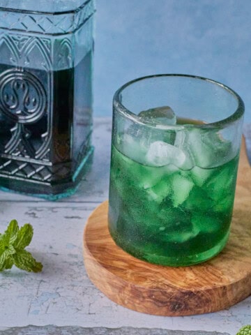 a glass of liqueur and ice on a round wooden board with a bottle of liqueur in the background, and a cork and some mint sprigs in the foreground