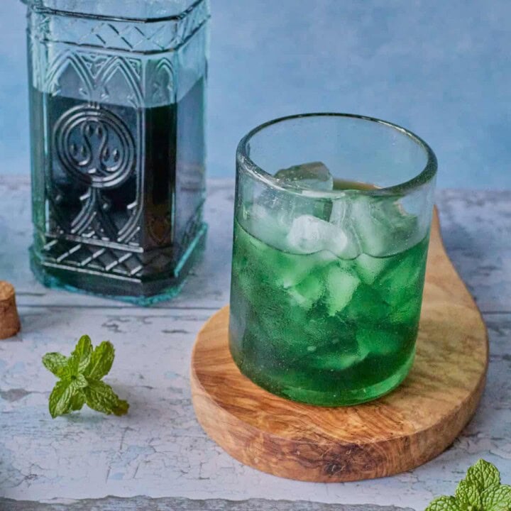 a glass of liqueur and ice on a round wooden board with a bottle of liqueur in the background, and a cork and some mint sprigs in the foreground