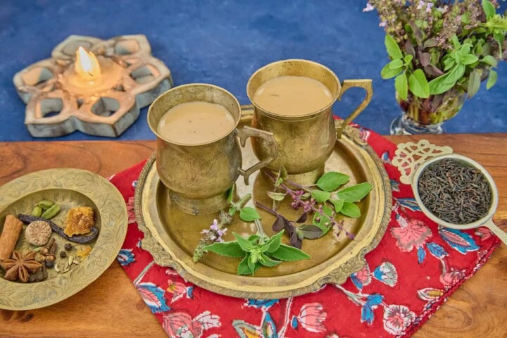 a brass teapot with two brass teacups along with 5 types of tulsi leaves on a brass dish, sitting on a printed napkin on a wooden cutting board, with a brass dish holding spices and a brass strainer full of black tea leaves, and a lit lotus-shaped wood candle and a vase of tulsi sprigs in the background