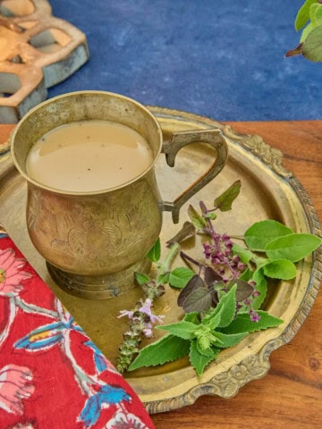 a square photo of a brass teacup along with 5 types of tulsi leaves on a brass dish, sitting on a printed napkin on a wooden cutting board, with a lit lotus-shaped wood candle and a vase of tulsi sprigs in the background