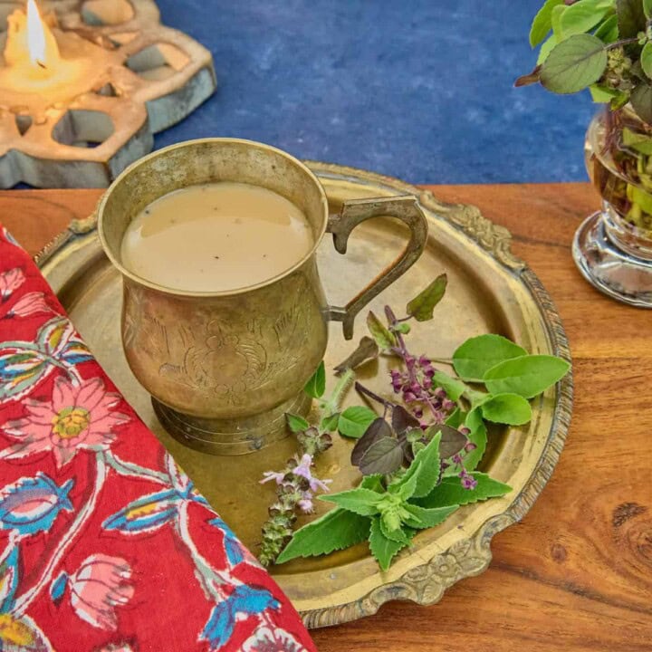 a square photo of a brass teacup along with 5 types of tulsi leaves on a brass dish, sitting on a printed napkin on a wooden cutting board, with a lit lotus-shaped wood candle and a vase of tulsi sprigs in the background