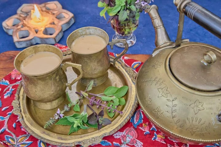 a brass teapot with two brass teacups along with 5 types of tulsi leaves on a brass dish, sitting on a printed napkin on a wooden cutting board, with a lit lotus-shaped wood candle and a vase of tulsi sprigs in the background