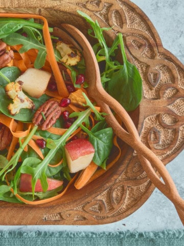 a square close-up photo of salad in a large carved wooden bowl with wooden mixing spoons and a napkin alongside