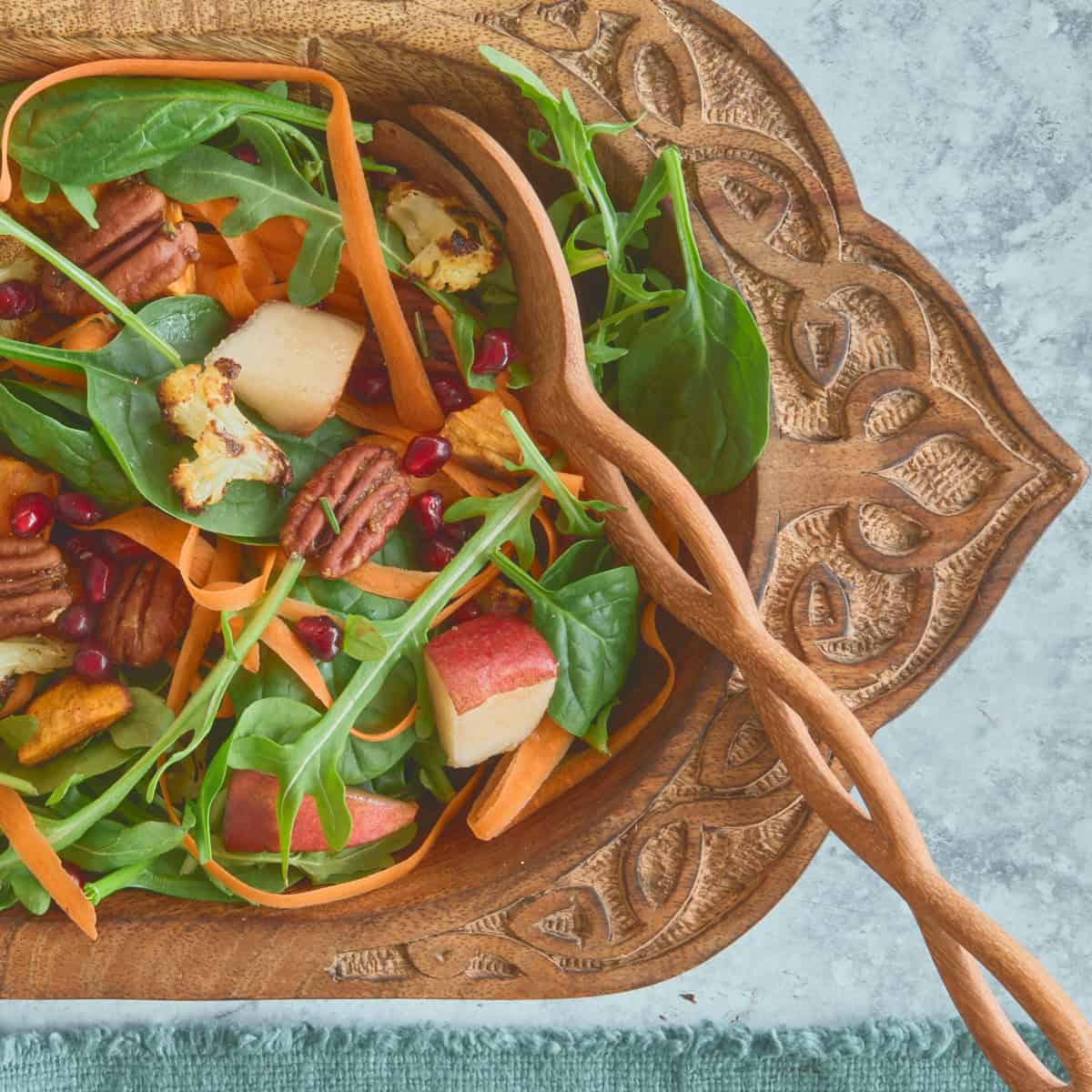 a square close-up photo of salad in a large carved wooden bowl with wooden mixing spoons and a napkin alongside