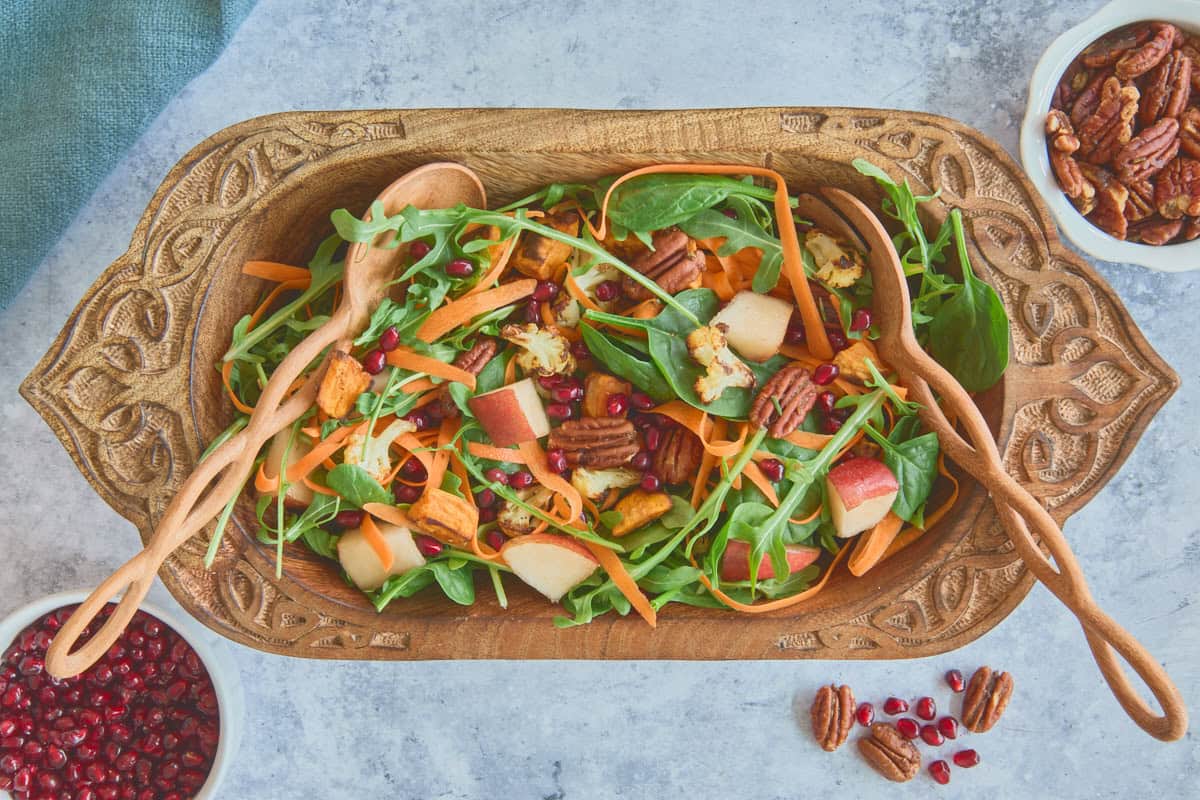 salad in a large oblate carved wooden bowl with wooden mixing spoons, and a napkin, bowl of spiced pecans, extra pecans and pomegranate arils, and a bowl of pomegranate arils alongside