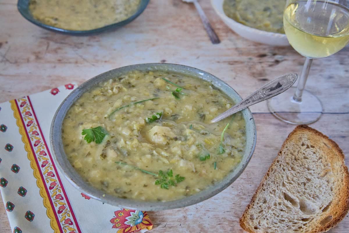 a bowl of soup, in which a spoon has been dipped, with a napkin, piece of toasted bread, glass of white wine, and two more bowls of soup and a spoon in the background