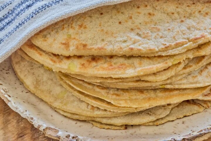 close-up photo of a stack of tortillas on a plate with a kitchen towel draped diagonally over the top