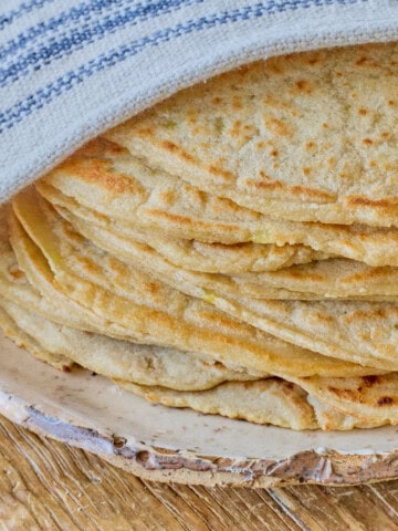 close-up square photo of a stack of tortillas atop a plate with a kitchen towel draped over the top