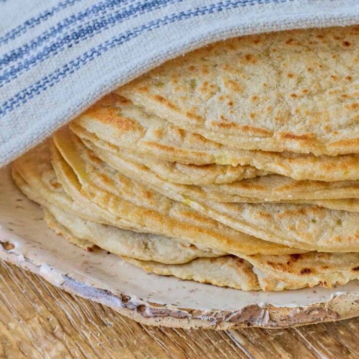 close-up square photo of a stack of tortillas atop a plate with a kitchen towel draped over the top