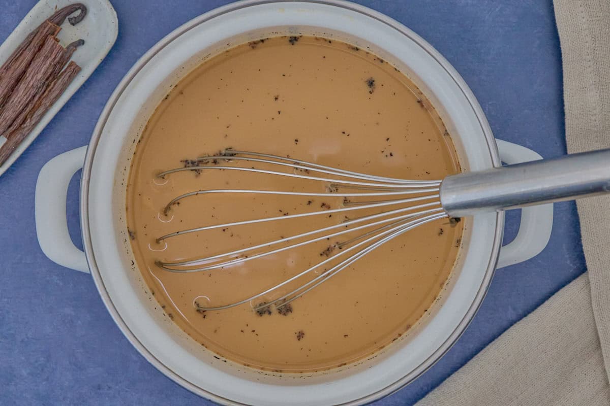 a pot with chai, with a metal whisk in the pot, and a dish of vanilla beans and kitchen towel in the background