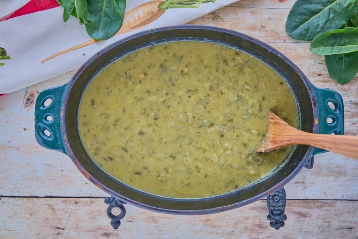 a cast iron pot filled with finished sorrel soup which is being stirred with a wooden spoon, with a kitchen towel and fresh sorrel leaves in the background
