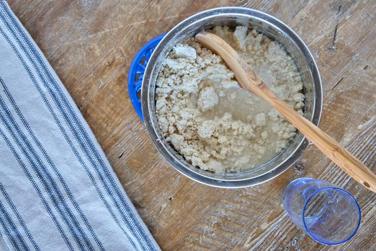 a bowl with water and dough ingredients being mixed together with a wooden spoon, and a kitchen towel and water glass on the side